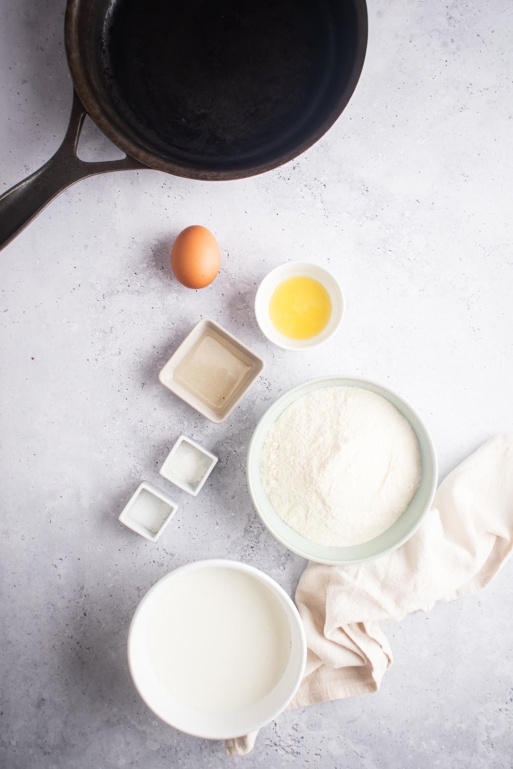 A egg, a bowl of melted butter, a bowl of flour, and a bowl of buttermilk all on a white counter.