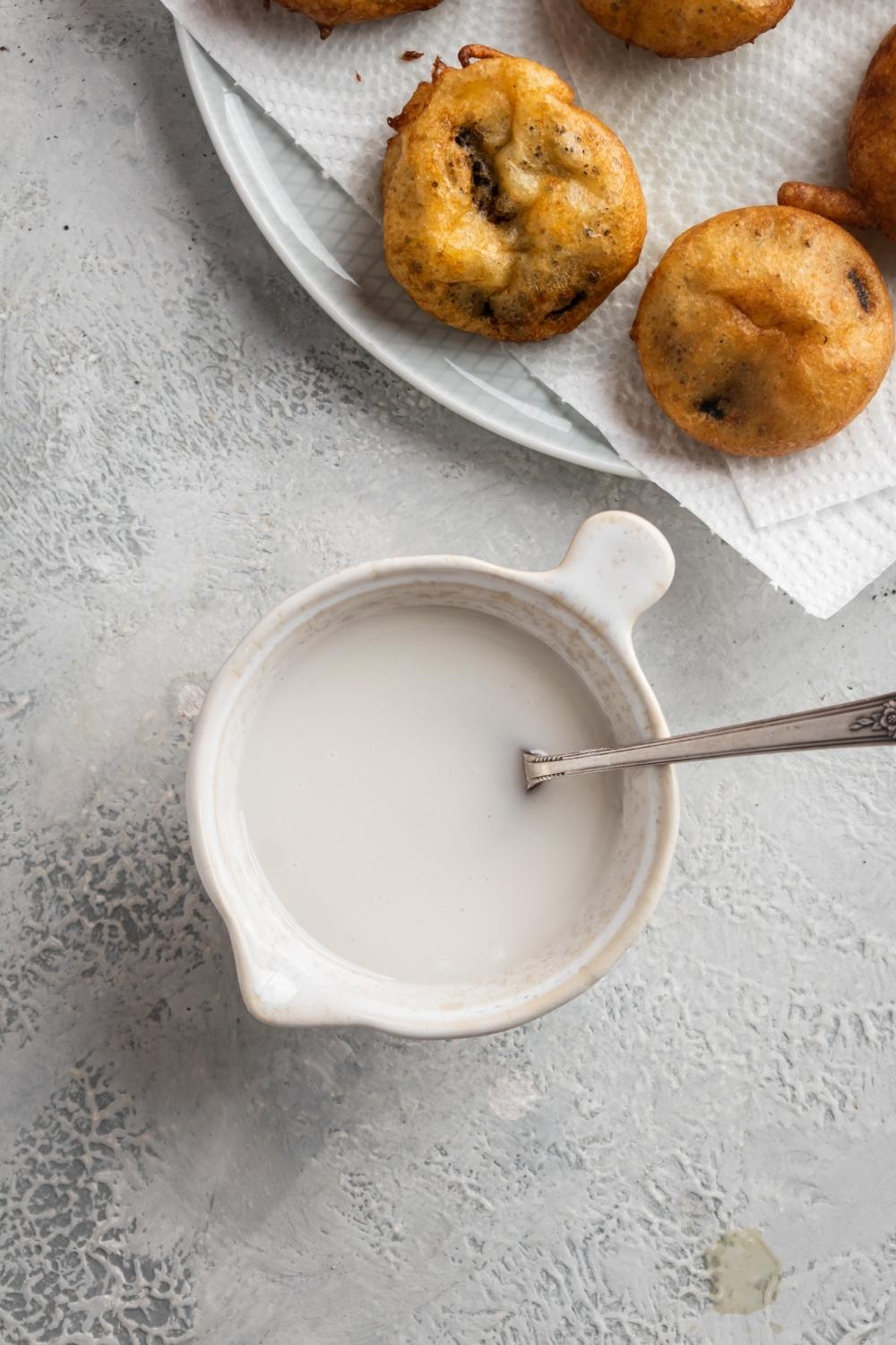 A ceramic bowl of glaze with a spoon in it. Above that is a plate with paper towels lining it and fried Oreos laid on top.