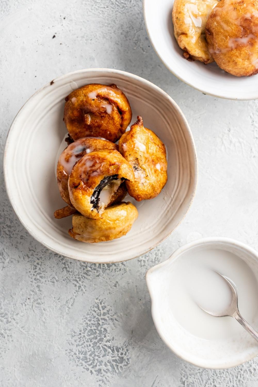 An overhead shot of glazed fried Oreos in a ceramic bowl on a countertop. The top fried Oreo has a bite out of it. To the right of the ceramic bowl with fried Oreos there is a bowl of glaze with a spoon in it.