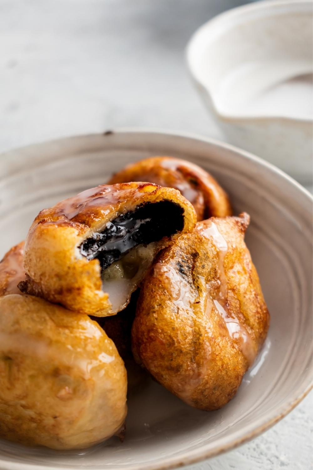 A pile of glazed fried Oreos in a ceramic bowl on the counter top. The top fried Oreo has a bite out of it.