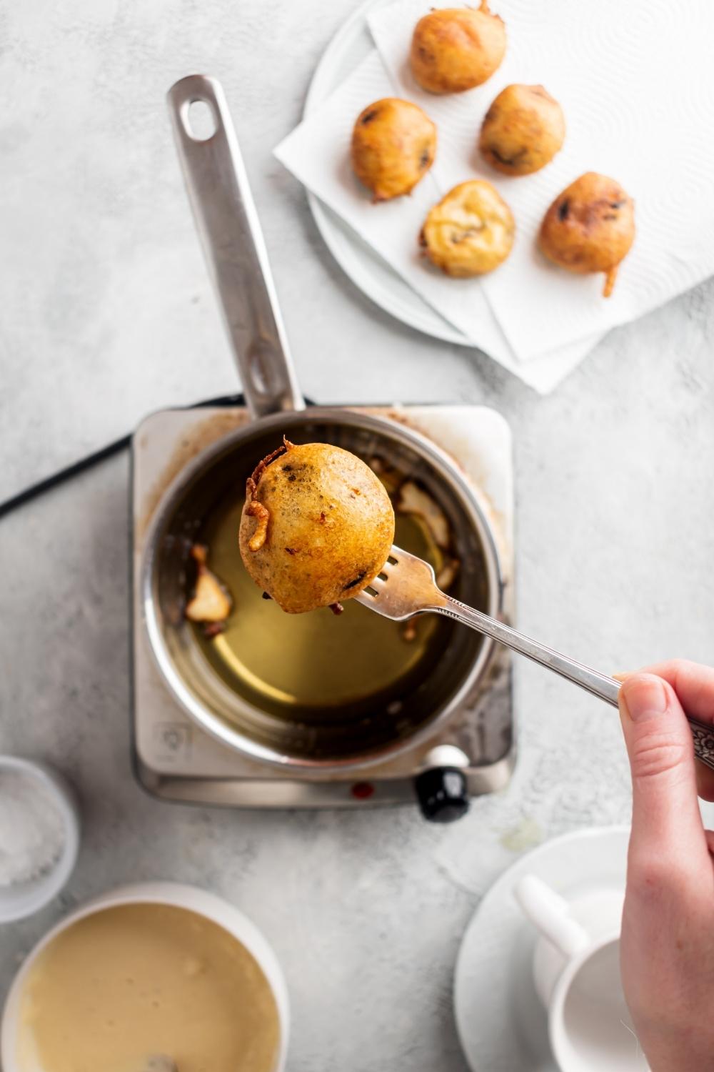 One fried Oreo on a fork above a pot of hot oil. Next to the pot of oil is a plate lined with paper towels with fried Oreos on top.