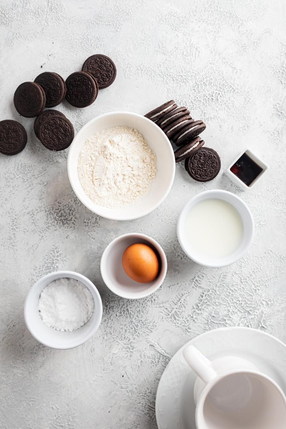 A countertop with one bowl of pancake powder mix, a bowl of milk, a bowl with an egg in it, a bowl with powdered sugar, and a bowl of vanilla extract. Oreos surround the bowls.