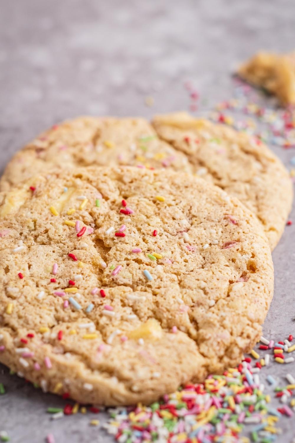 Two funfetti cookies on a parchment paper sheet. Rainbow sprinkles are on top of the cookies and next t them.
