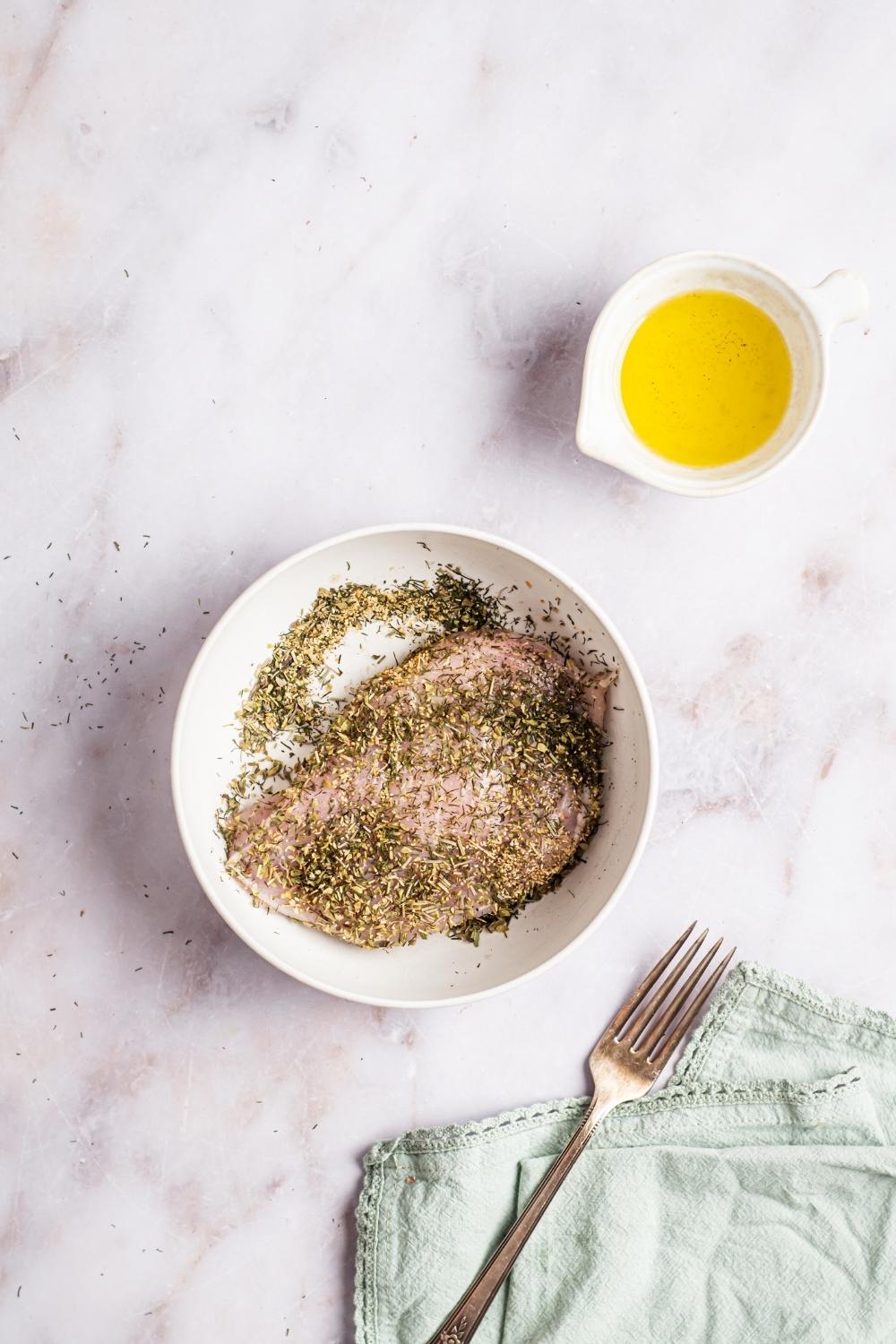 Chicken covered in herb seasoning in a white bowl. A bowl of olive oil is behind it.