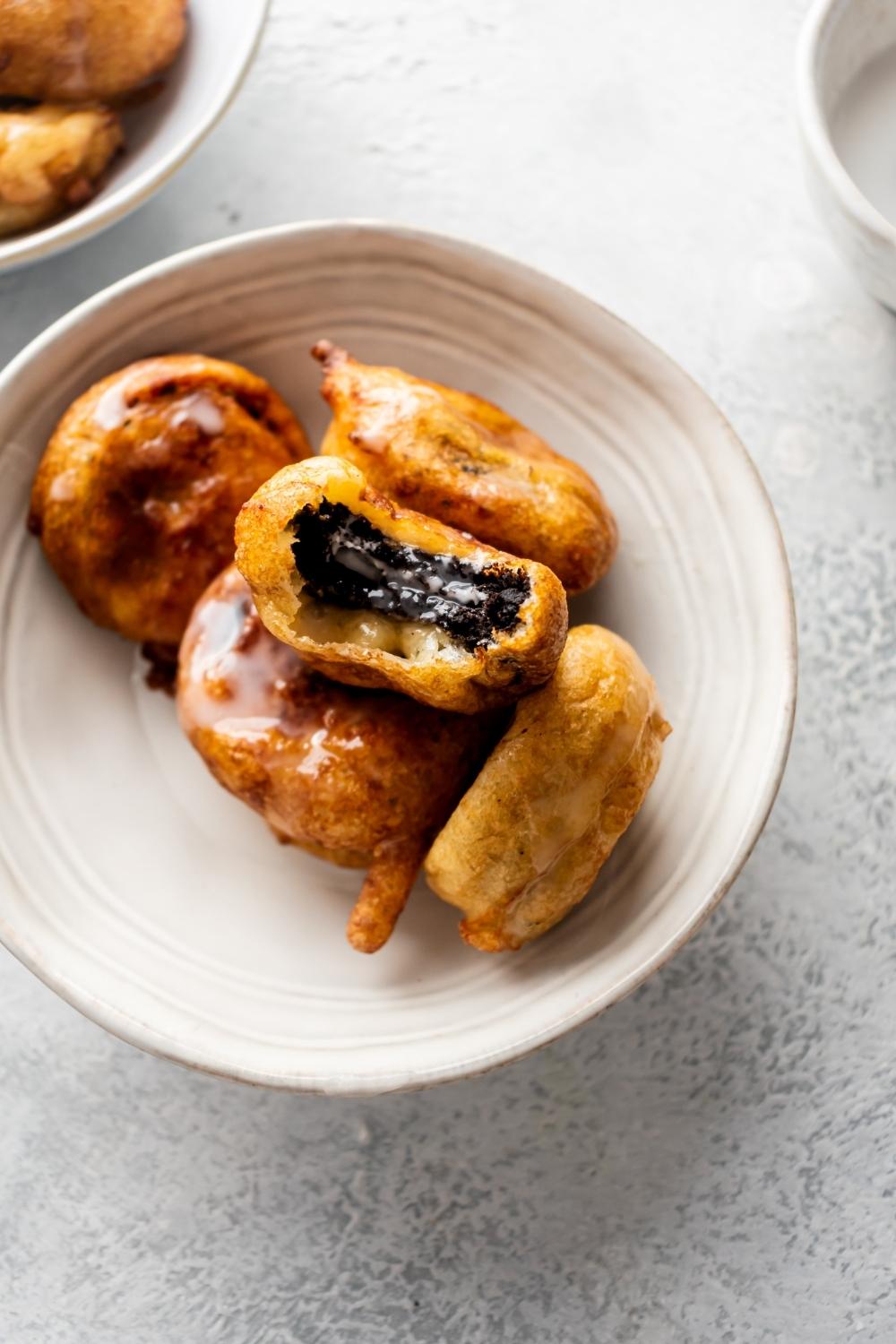 A pile of fried oreos with glaze. One oreo with a bite out of it in a ceramic bowl on a countertop