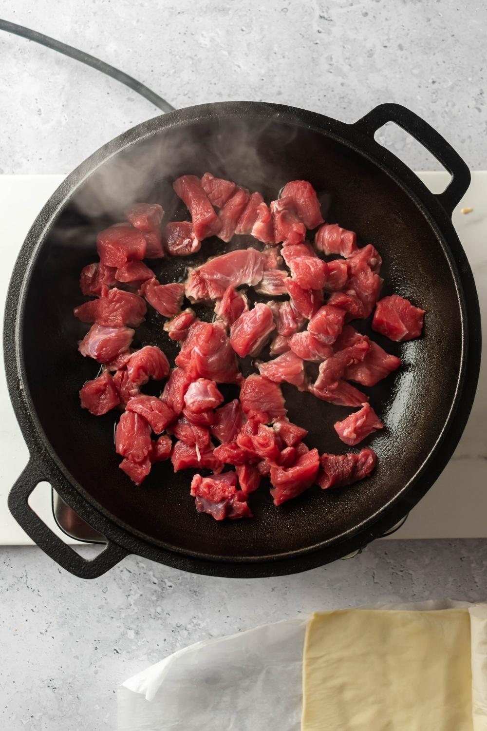 An overhead view of raw beef chunks being cooked in an iron pot.