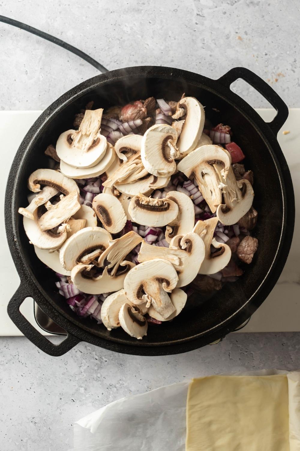 An overhead view of an iron pot with beef chunks, diced onions, and sliced mushrooms.