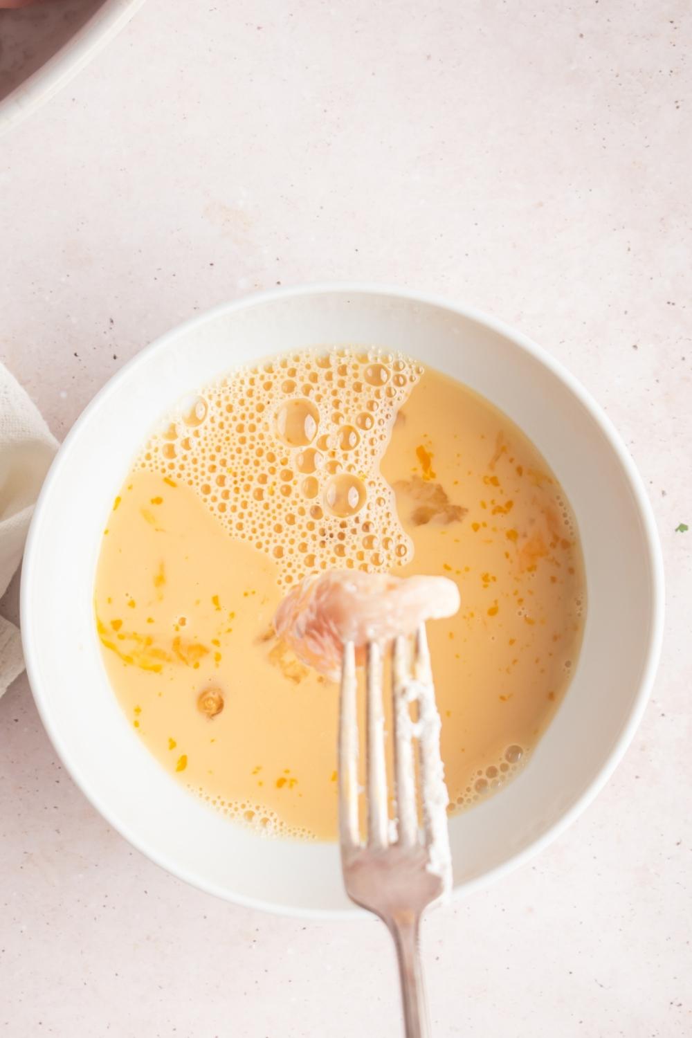 And over a deal of the egg batter in a bowl. A fork holds an uncooked chicken chunk over the bowl.