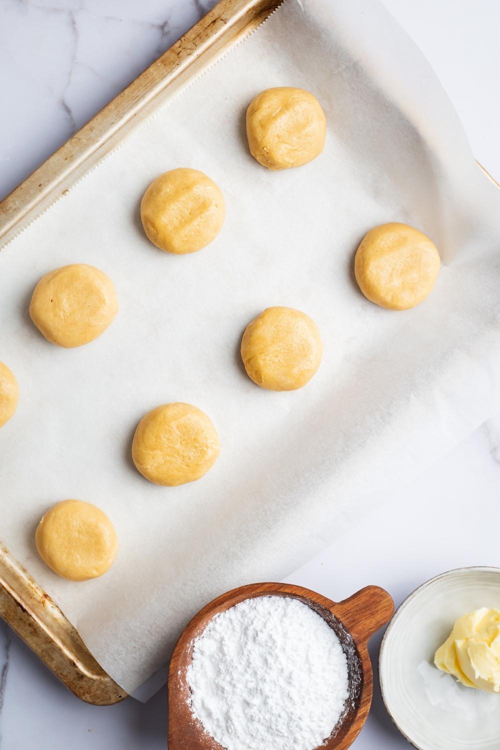 An overhead view of a baking sheet lined with parchment paper. There are eight flattened balls of crumbl cookie dough on it.