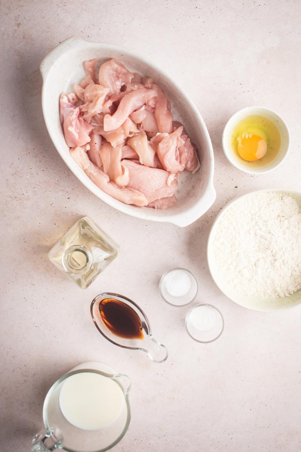 An overhead view of a casserole dish containing cut up uncooked chicken. There are multiple balls surrounding the casserole dish containing an egg, flour, soy sauce, milk, salt, and baking powder.