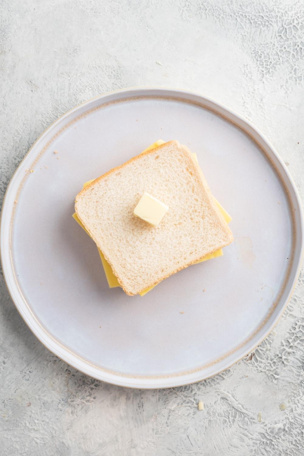 An overhead view of a plate with two white bread slices with cheese in the middle of them and a square of butter on top of them.