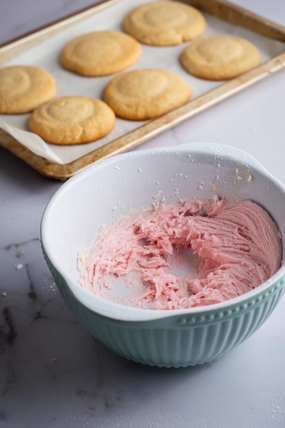 A large bowl sits on a countertop with pink icing. There is a baking sheet lined with parchment paper holding six sugar cookies.