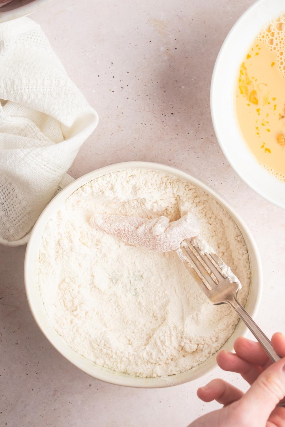 An overhead view of a fork dunking an uncooked chicken chunk in flour.