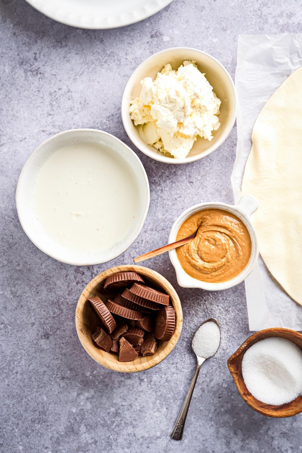 An overview of all of the Reeses pie ingredients. A large bowl holds heavy whipping cream, a smaller bowl holds cream cheese, the same size bowl holds Reeses peanut butter cups, a smaller bowl holds creamy peanut butter, and a small bowl next to that holds sugar.