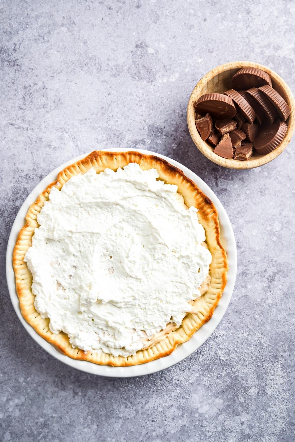 An overhead view of a Reeses pie topped with homemade whipped cream and a small bowl sits next to that with Reese's peanut butter cups.