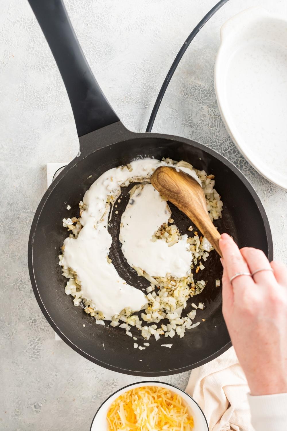 An overhead view of a skillet with a hand holding a wooden spoon stirring cream into the cooked garlic and onion mixture.