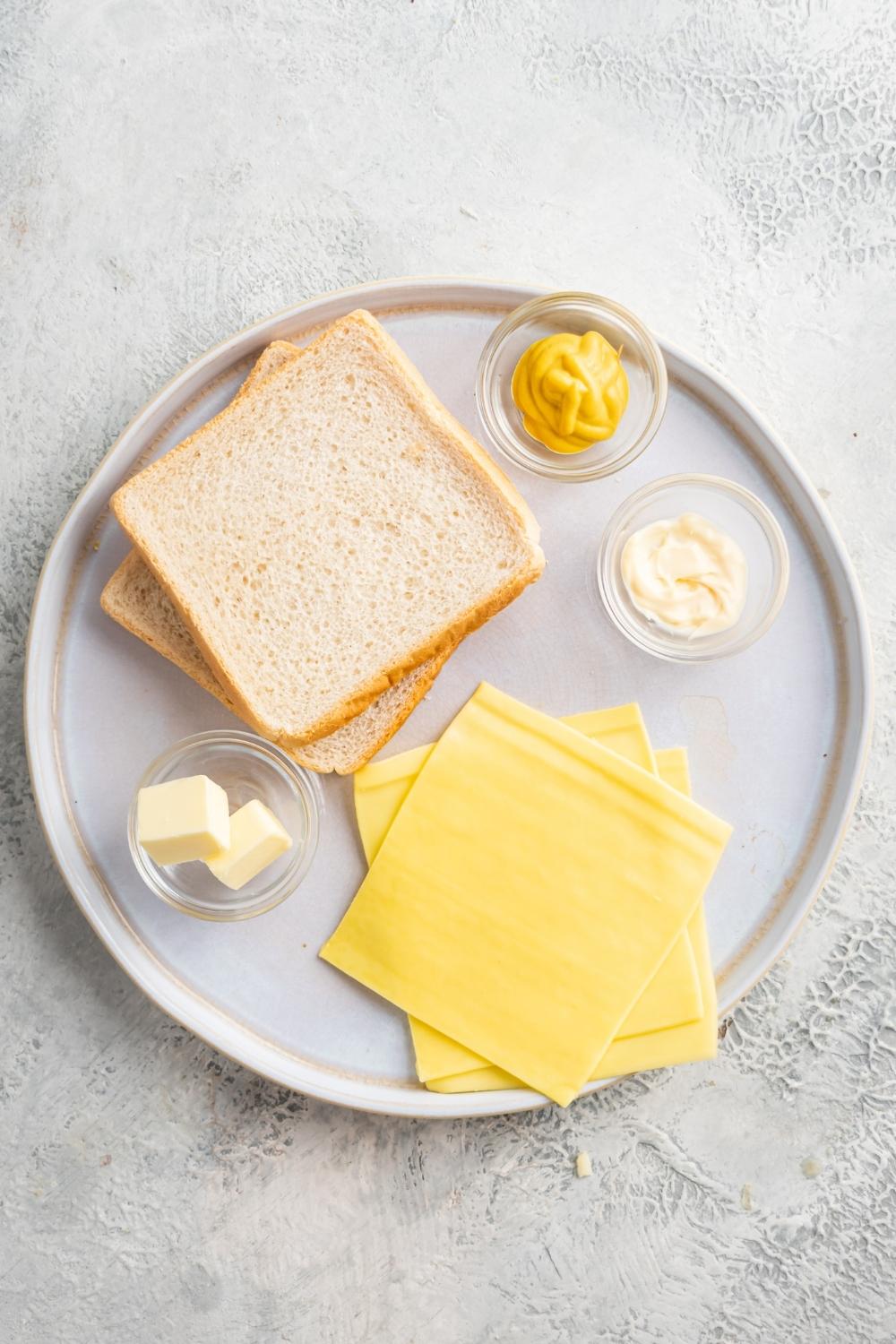 An overhead view of two slices of white bread, three slices of cheese, and three small bowls containing mustard, mayo, and butter.