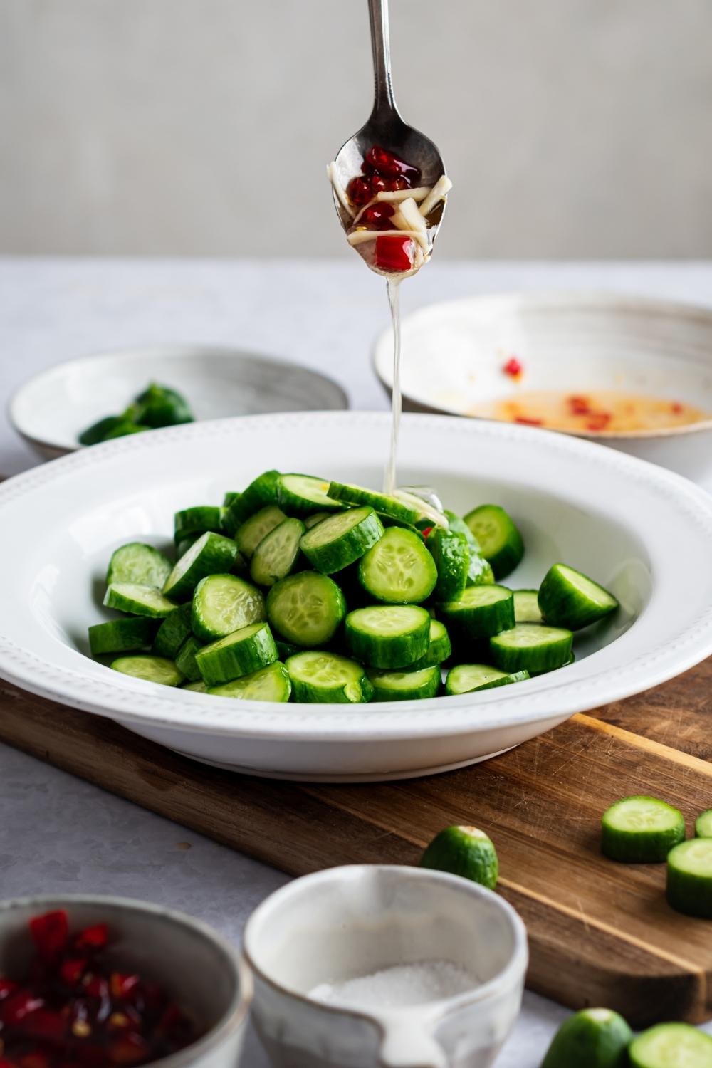 A spoon pouring a dressing on top of diced cucumbers in a white bowl on top of a wooden cutting board.