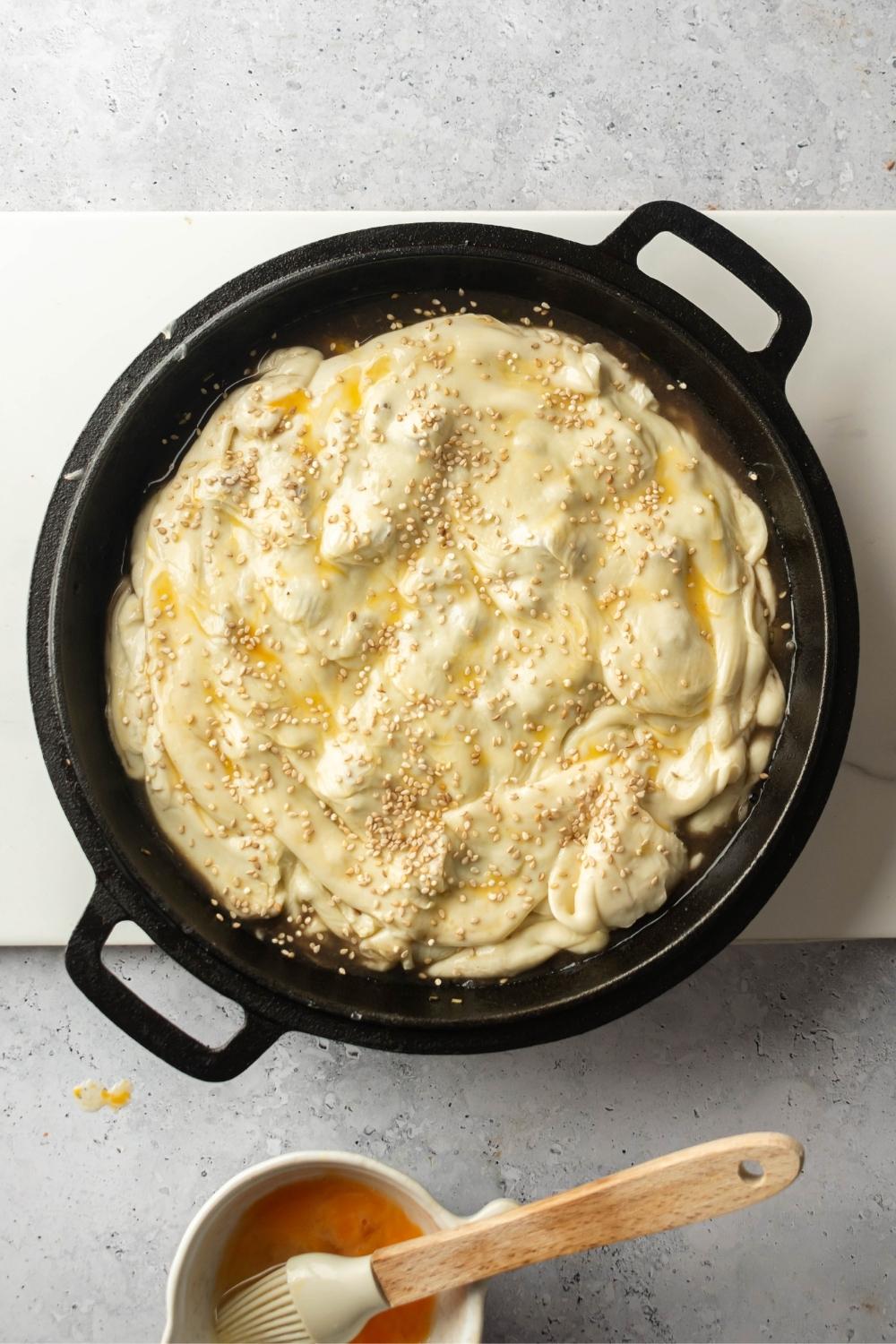 An overhead view of an iron pot containing beef pot pie ingredients all covered with a raw pie crust. The pie crust has been egg washed and has sesame seeds on top.