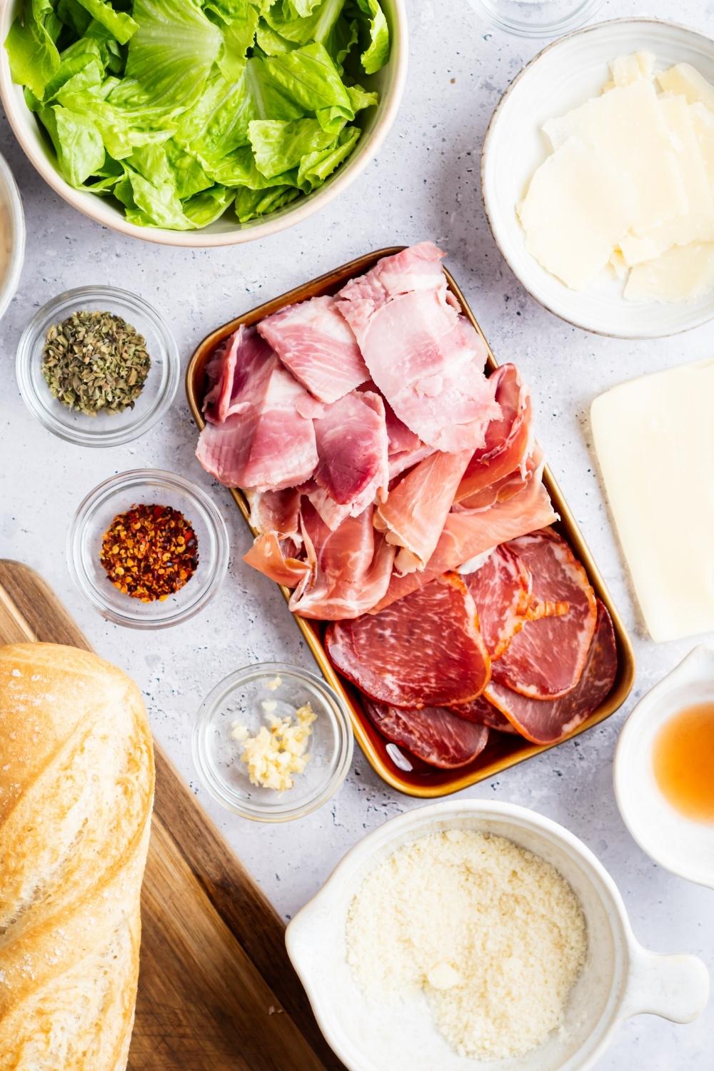 An overhead view of a tray of Italian cold cuts, a bowl of chopped Romain lettuce, a bowl of fresh shaved Parmesan, a bowl of grated Parmesan, and three small bowls containing red pepper flakes, minced garlic, oregano.