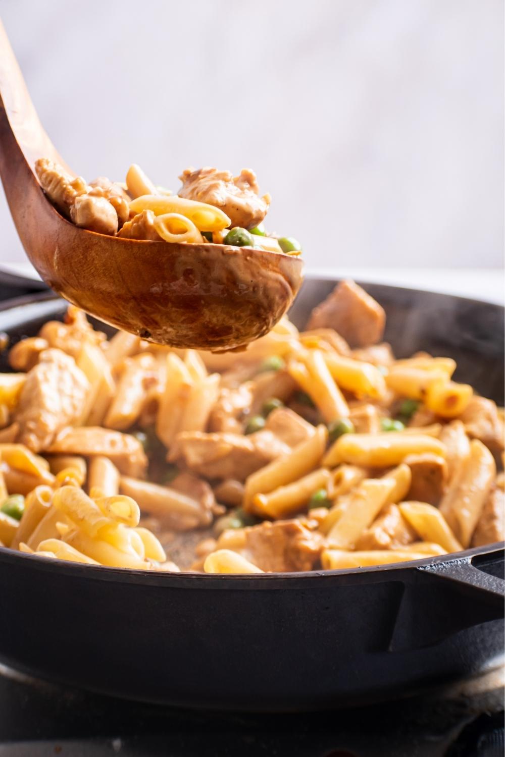 A wooden spoon with a scoop of Spicy Chicken Chipotle Pasta. A skillet is below full of the remaining Spicy Chicken Chipotle Pasta.