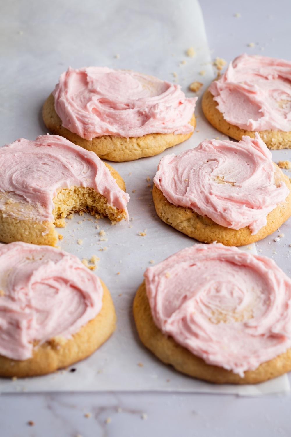 A close-up of six sugar cookies with pink icing on them laying on a parchment paper. One of the cookies has a bite taken out of it.