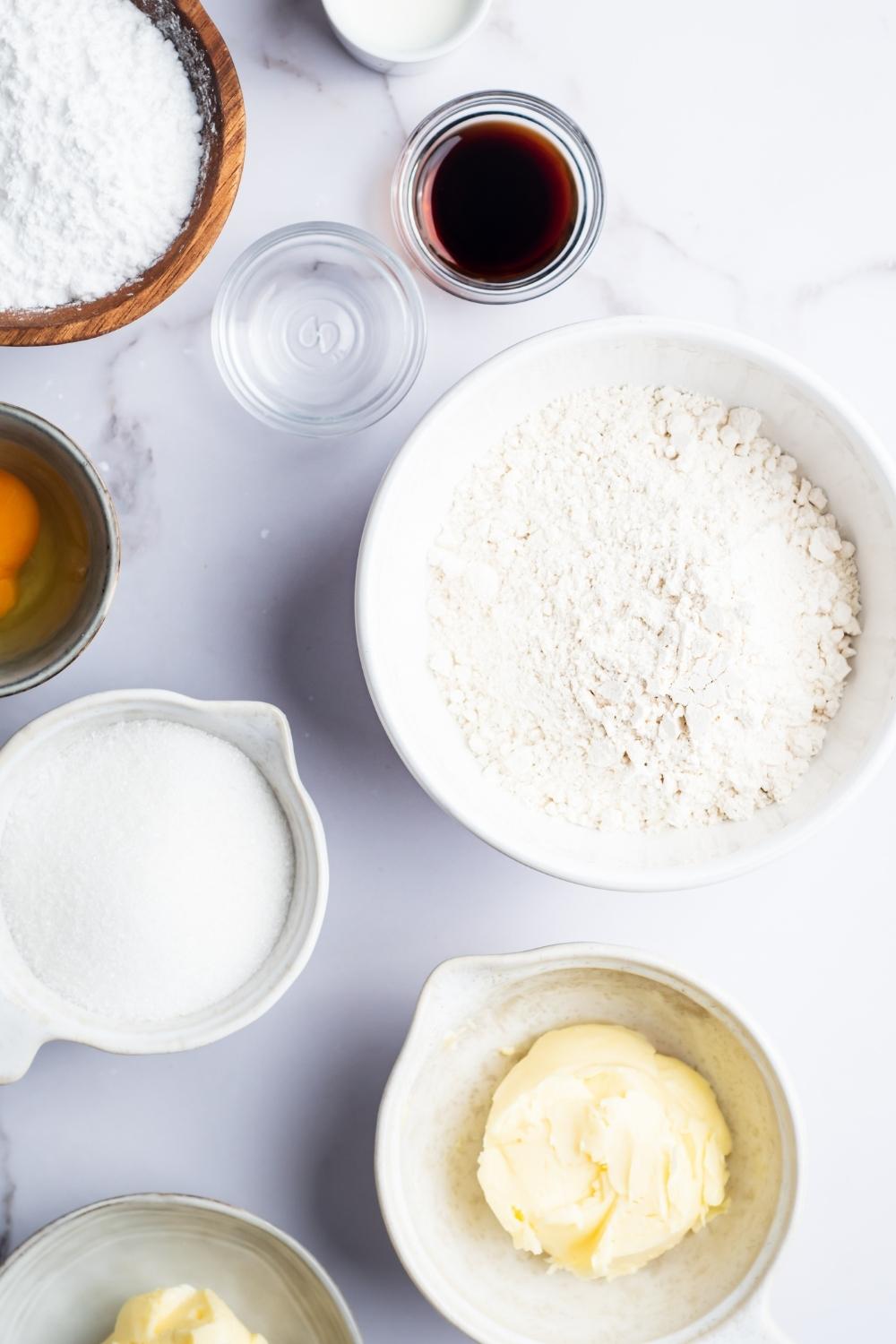 An overhead view of a large bowl with flour in it, a medium size bowl with powdered sugar, and a smaller bowl with granulated sugar and six small bowls with butter, egg, vanilla extract, almond extract, and milk