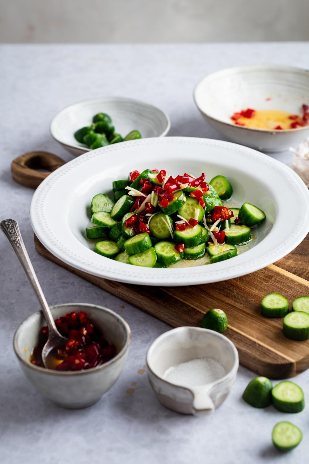 Diced chilis and cucumbers in a white bowl on top of a wooden cutting board. In front of it is a bowl of the chilis and a bowl of salt.