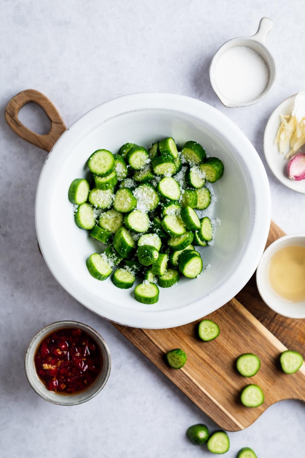 Sliced cucumbers with salt on top in a white bowl on top of a wooden cutting board.