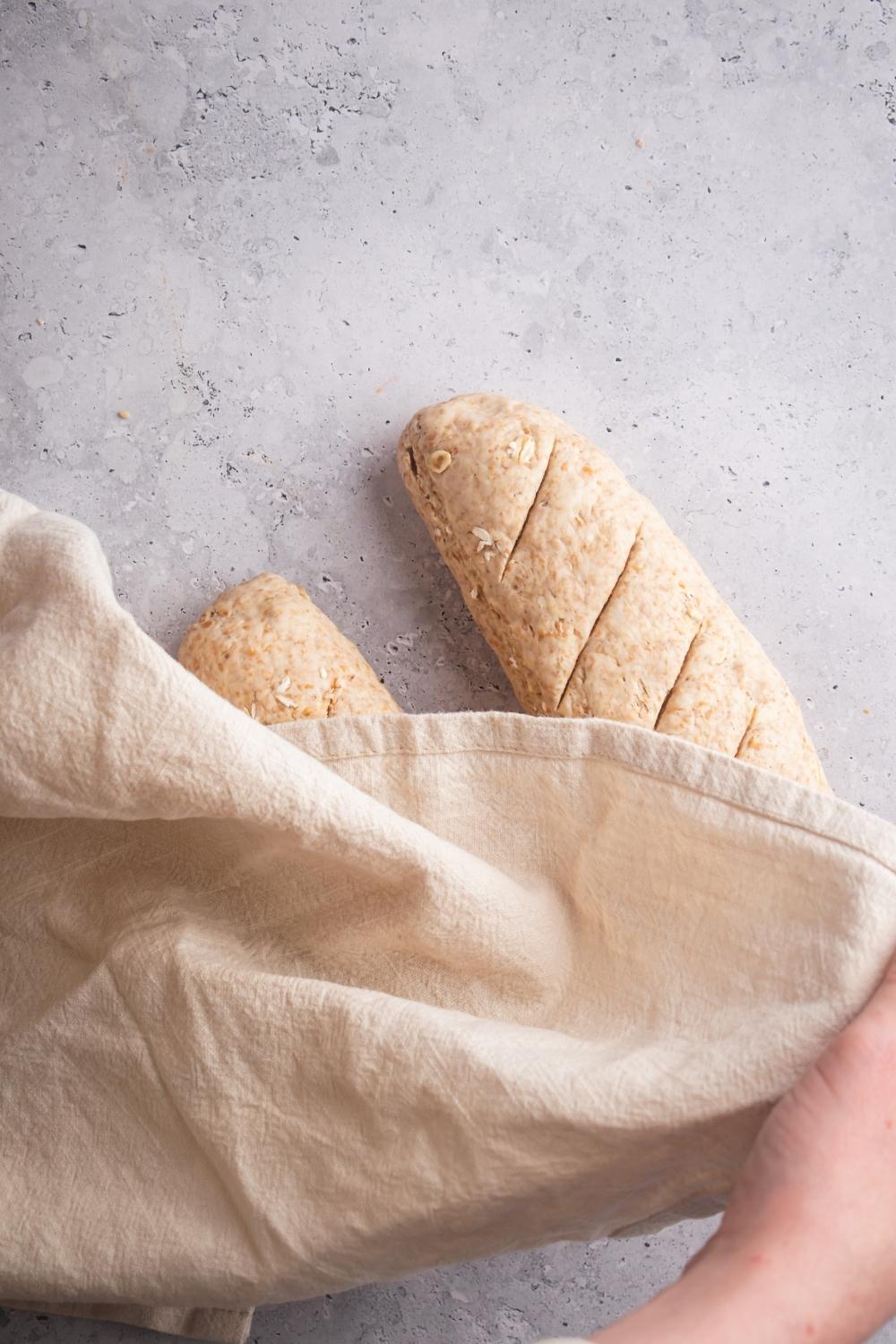 To unbaked bread those made in two loaves with slits on the top. A hand covers both of the uncooked loaves with a kitchen towel.