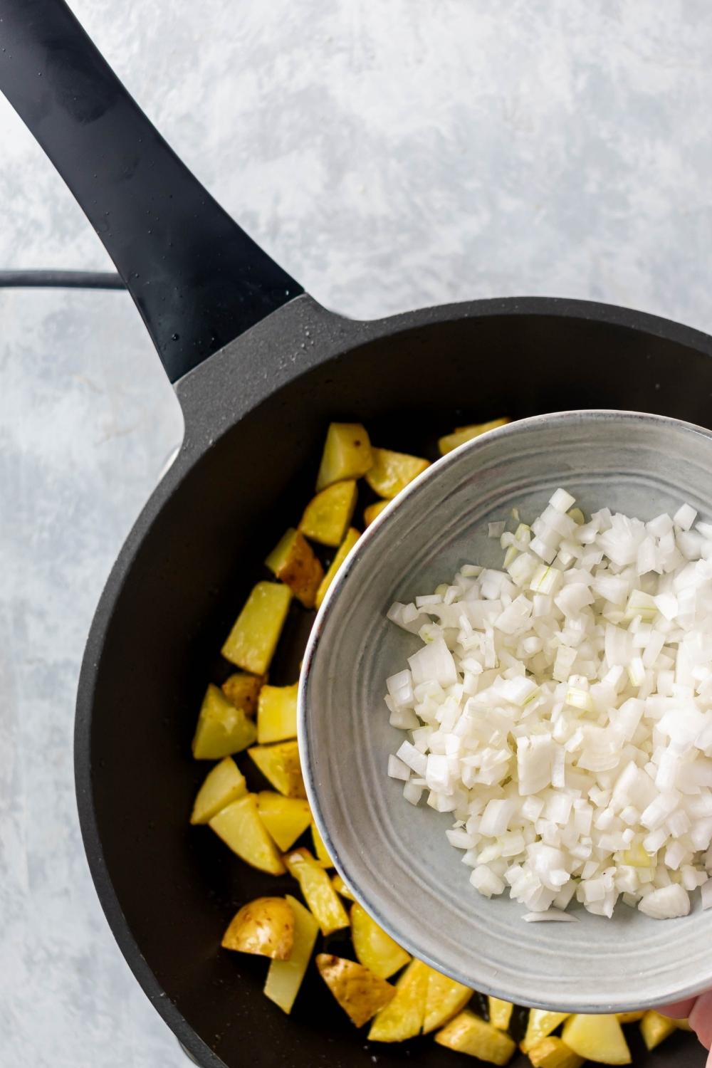 An overhead view of an iron pan with cubed potatoes. A small bowl with diced onions is overtop the pan.