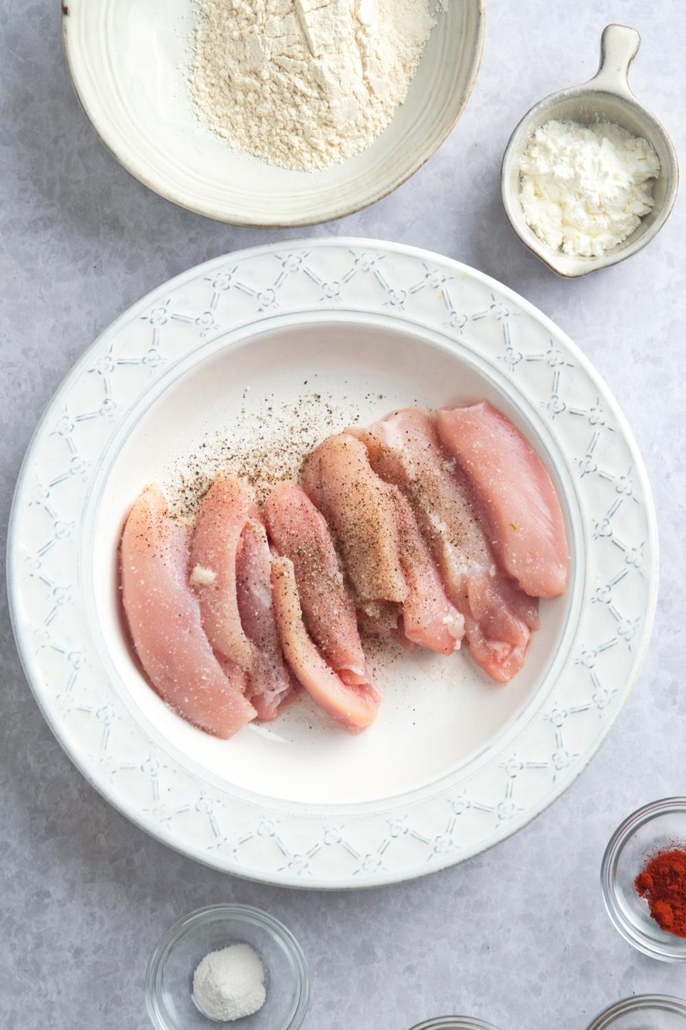 An overhead seal of raw chicken on a white dish seasoned with salt and pepper. A medium size bowl sits next to the dish with flour in it and a small bowl sits next to that bowl containing cornstarch.