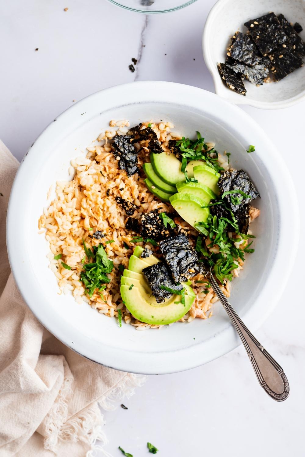 An overhead view of TikTok salmon rice bowl in a dish with a fork sitting in it. It is topped with avocado, cilantro, and seaweed snacks.