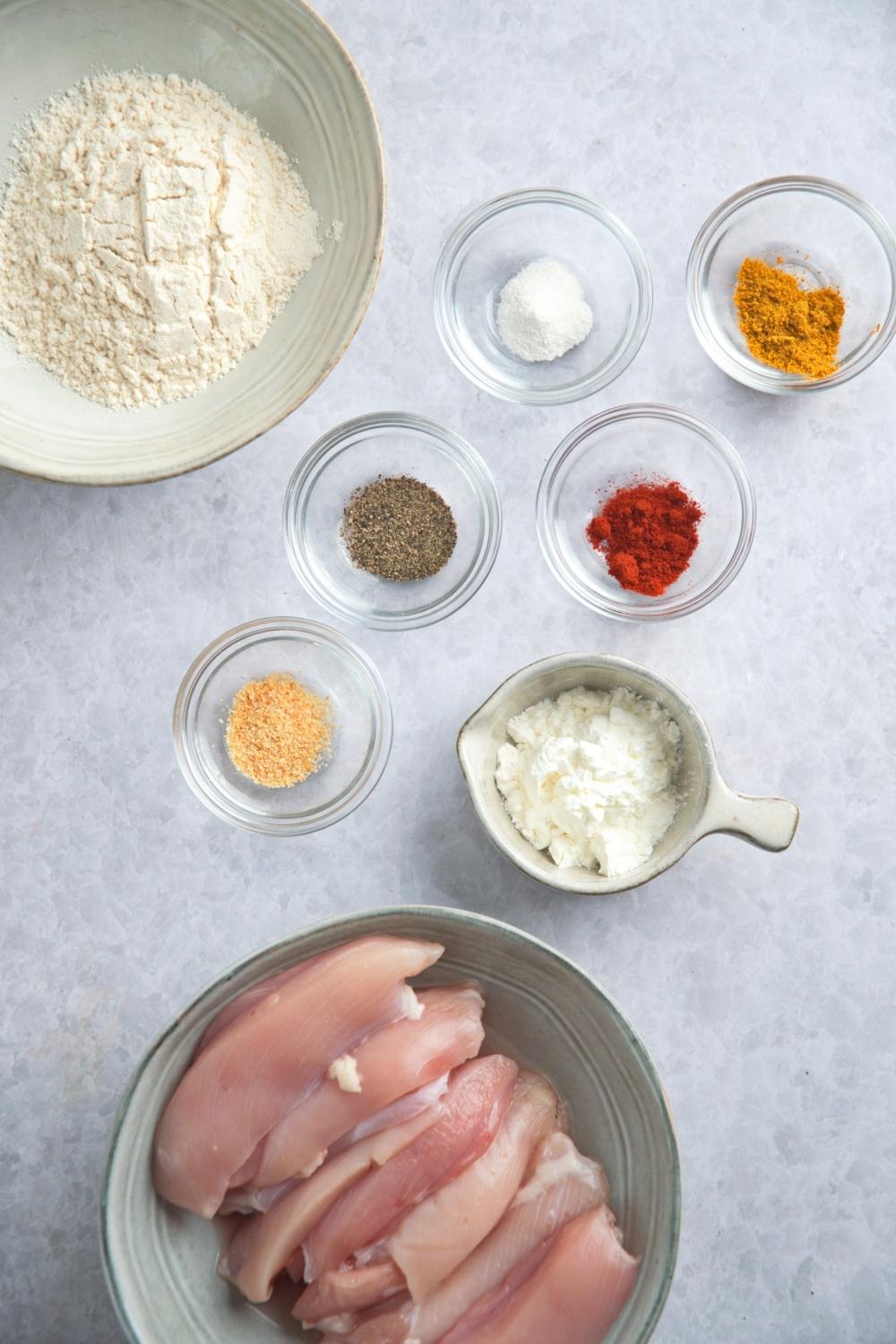 An overhead view of a large bowl containing raw chicken strips, a medium size bowl containing flour, and six small bowls containing spices and cornstarch.