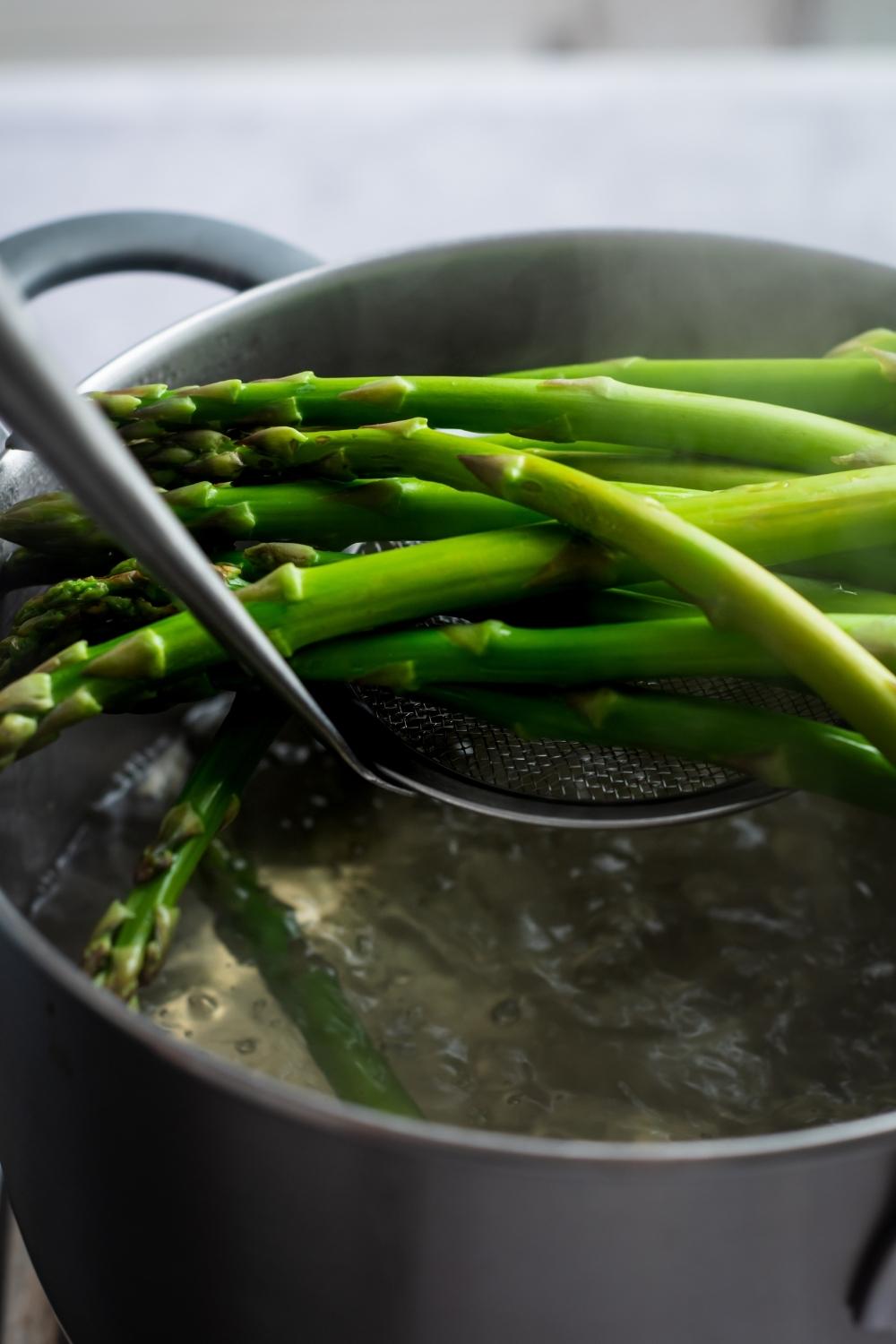 A deep pot with boiling water and a slottes spoon dishing out the bright green cooked asparagus.