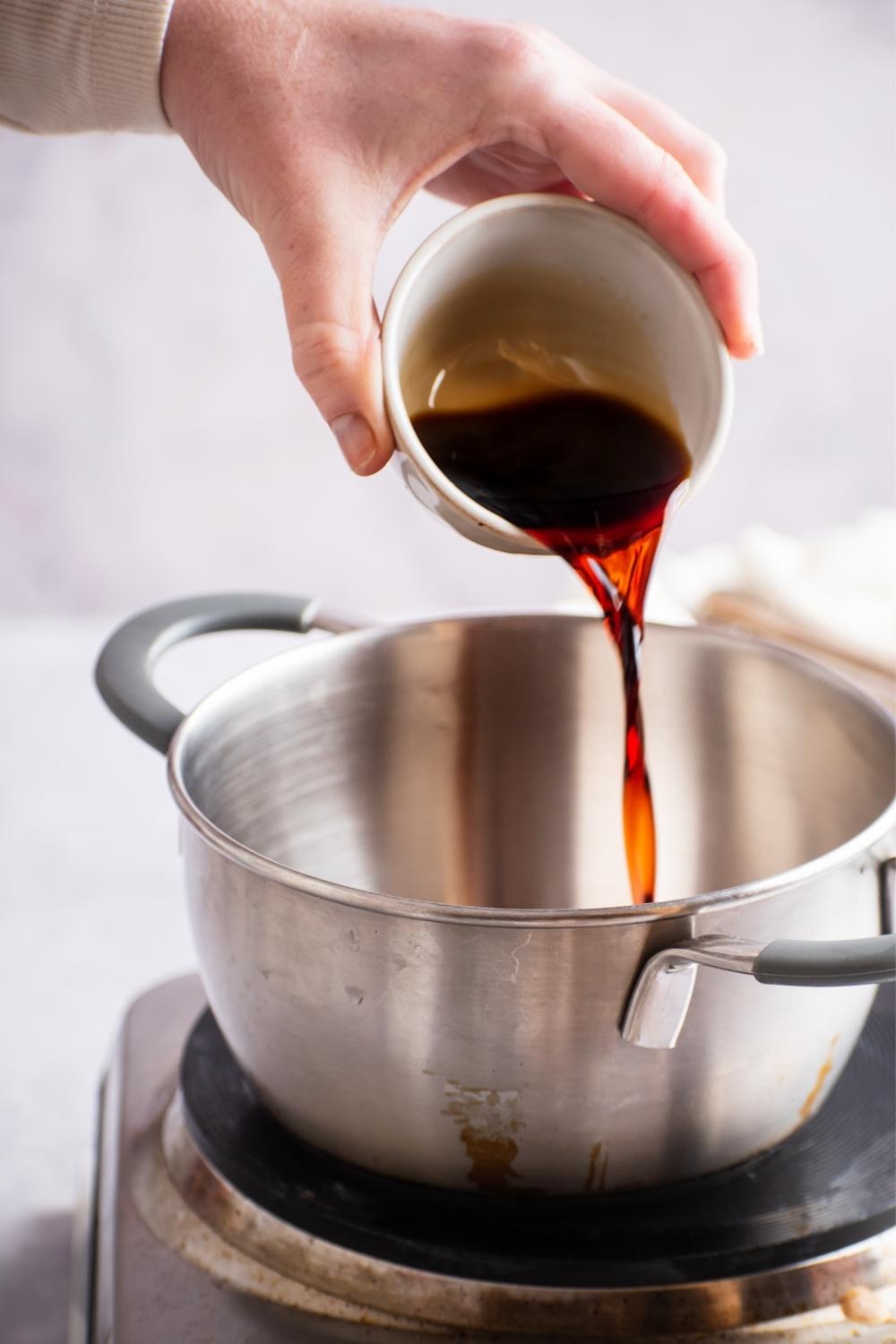A hand pouring soy sauce into a pot on a burner.