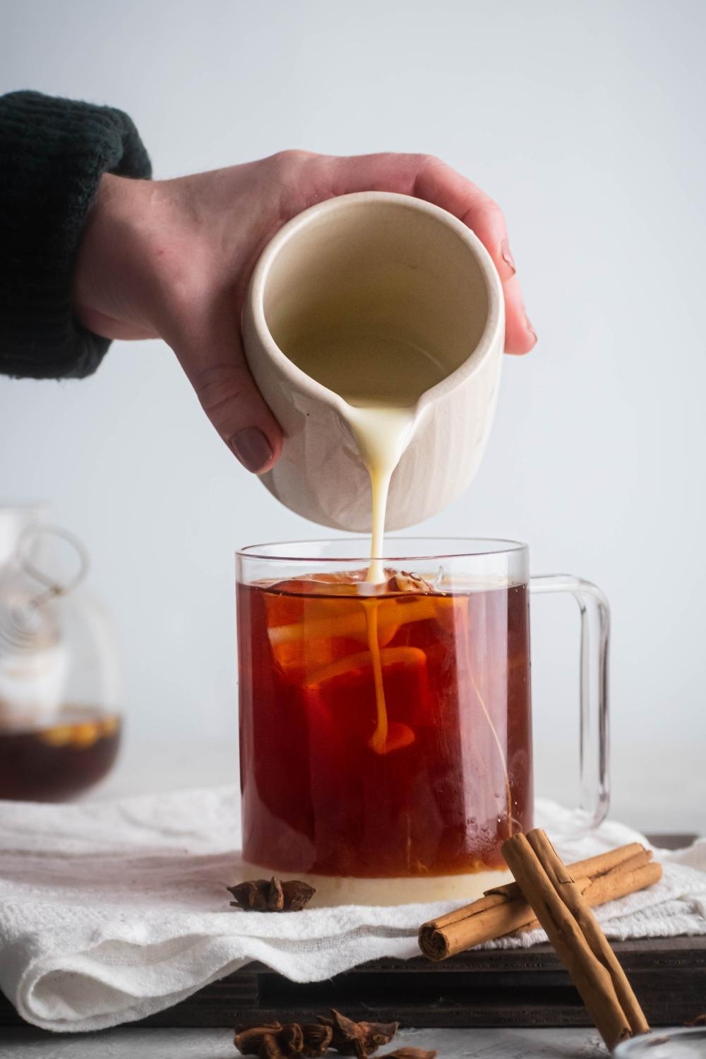 A hand pouring milk into a glass cup filled with black tea.