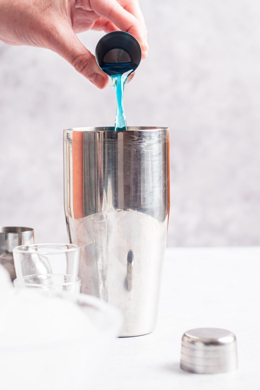 A hand using a jigger to pour blue curacao into a shaker. Two shot glasses sit next to the shaker and the shaker top is on the counter as well.