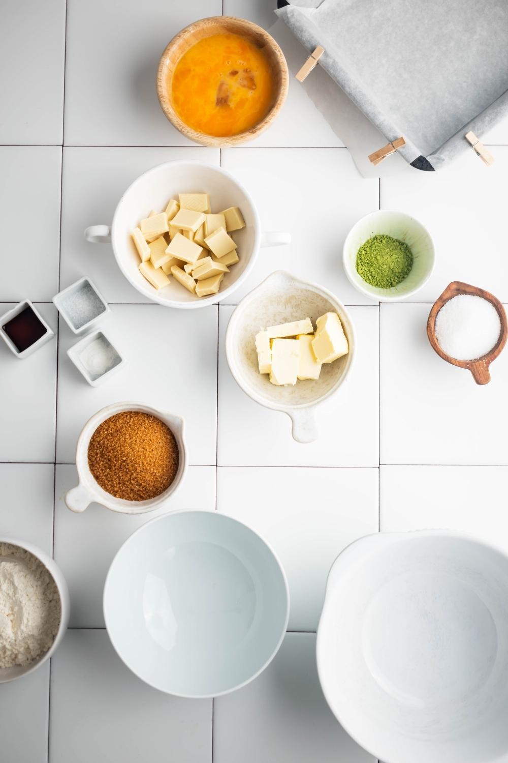 A bowl with whisked egg, a bowl with chopped butter, a bowl with matcha powder, and a bowl of brown sugar all on a white counter.