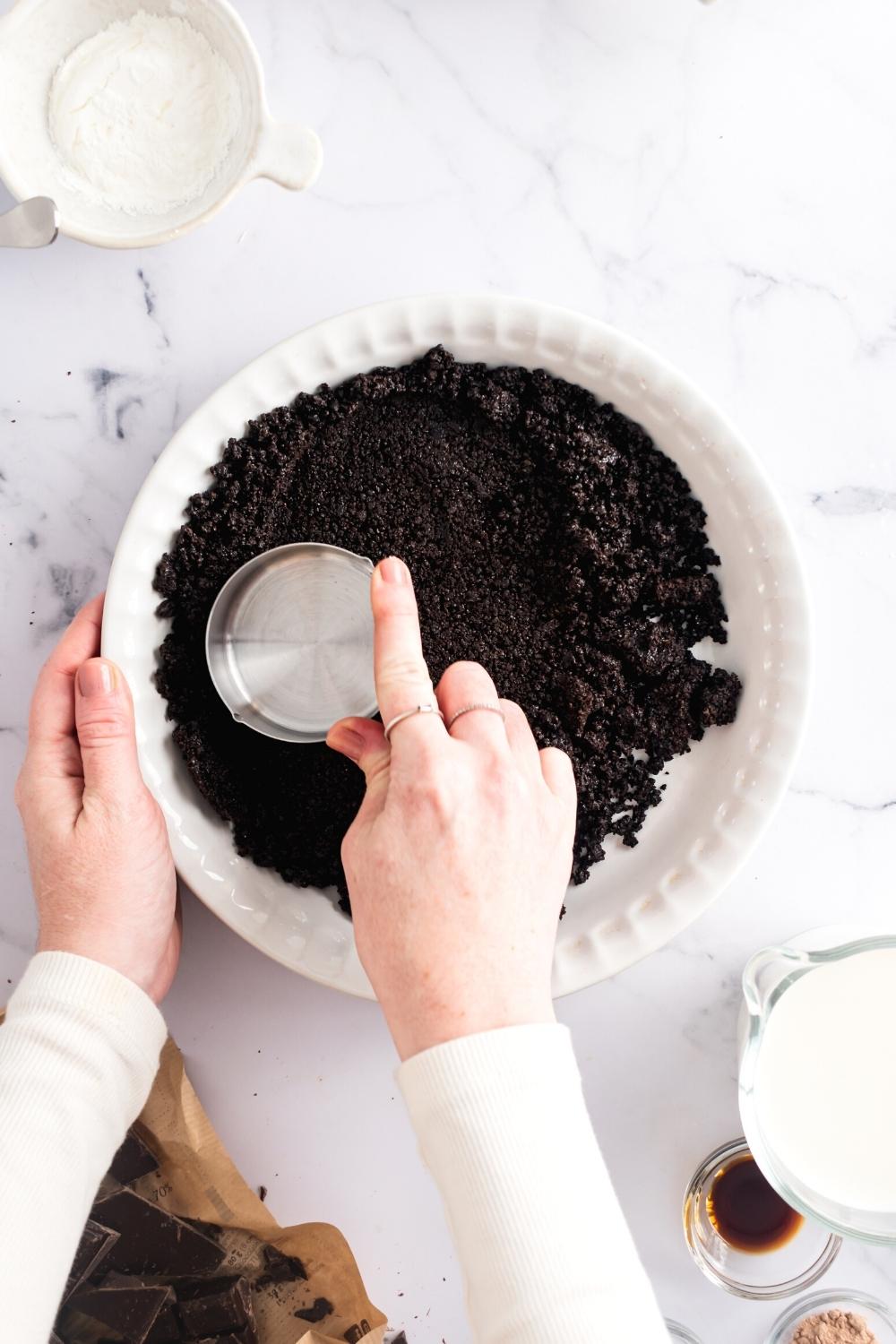 A hand holding a cup forming crushed Oreos into a pie dish.