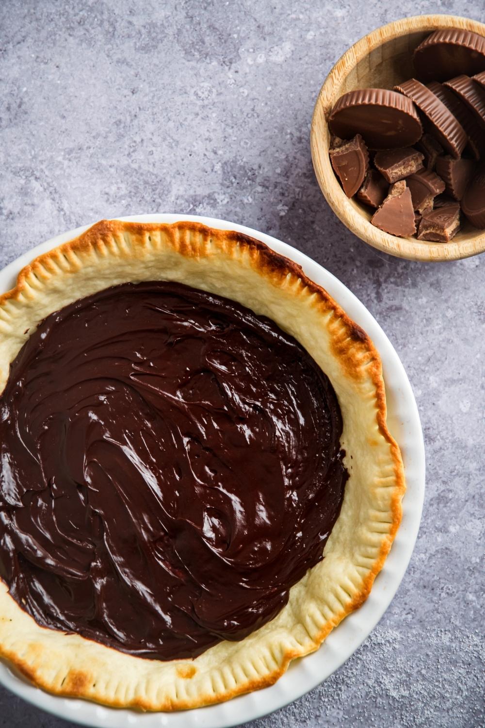An overview of a baked pie crust topped with chocolate fudge. A small wooden bowl sits next to it with Reese's peanut butter cups.