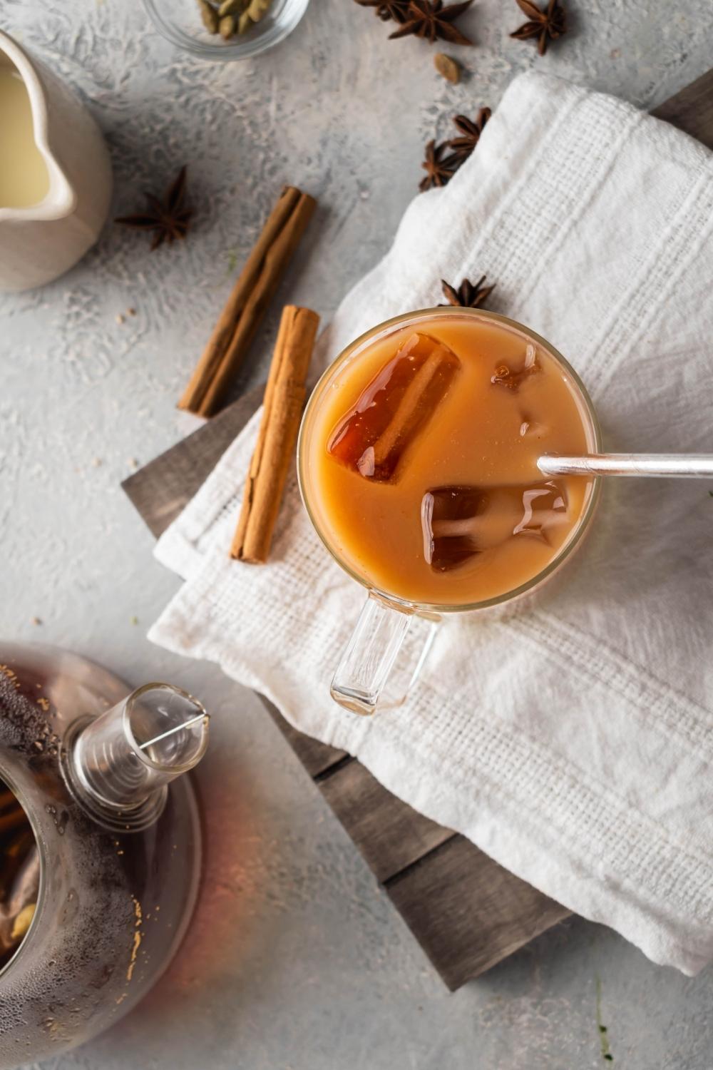 A glass on thai tea on top of a white napkin on a wood board on a grey counter. Next to it is part of a glass pitcher with the tea in it.