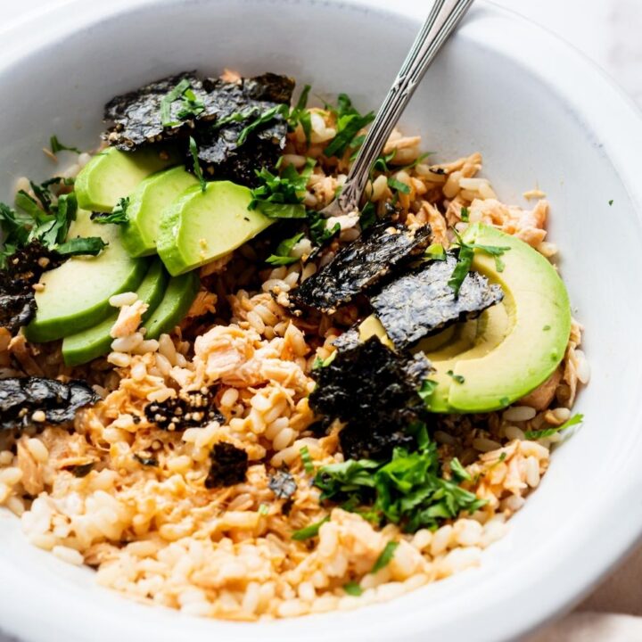 A close up of TikTok salmon rice bowl in a dish with a fork sitting in it. It is topped with avocado, cilantro, and seaweed snacks.