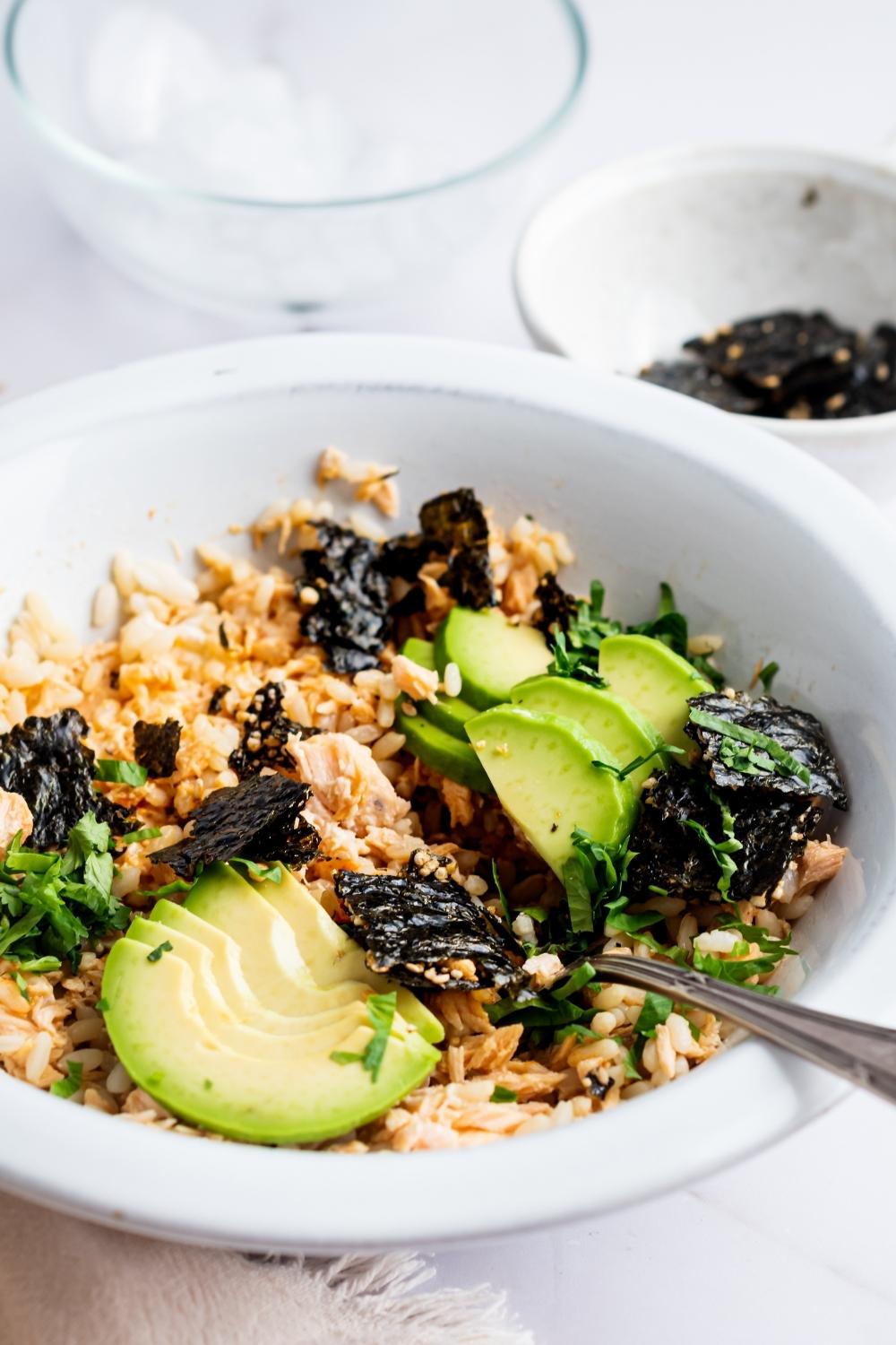TikTok salmon rice bowl in a dish with a fork sitting in it. It is topped with avocado, cilantro, and seaweed snackers