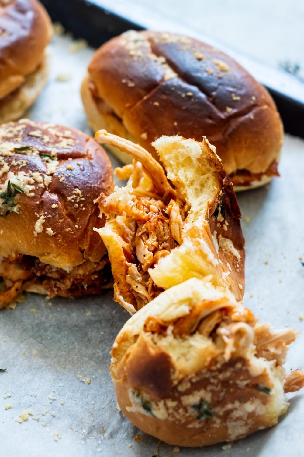 A close-up of BBQ shredded chicken sliders on a parchment lined baking tray. One of the barbecue chicken sliders is cut in half to reveal the chicken between the bread.
