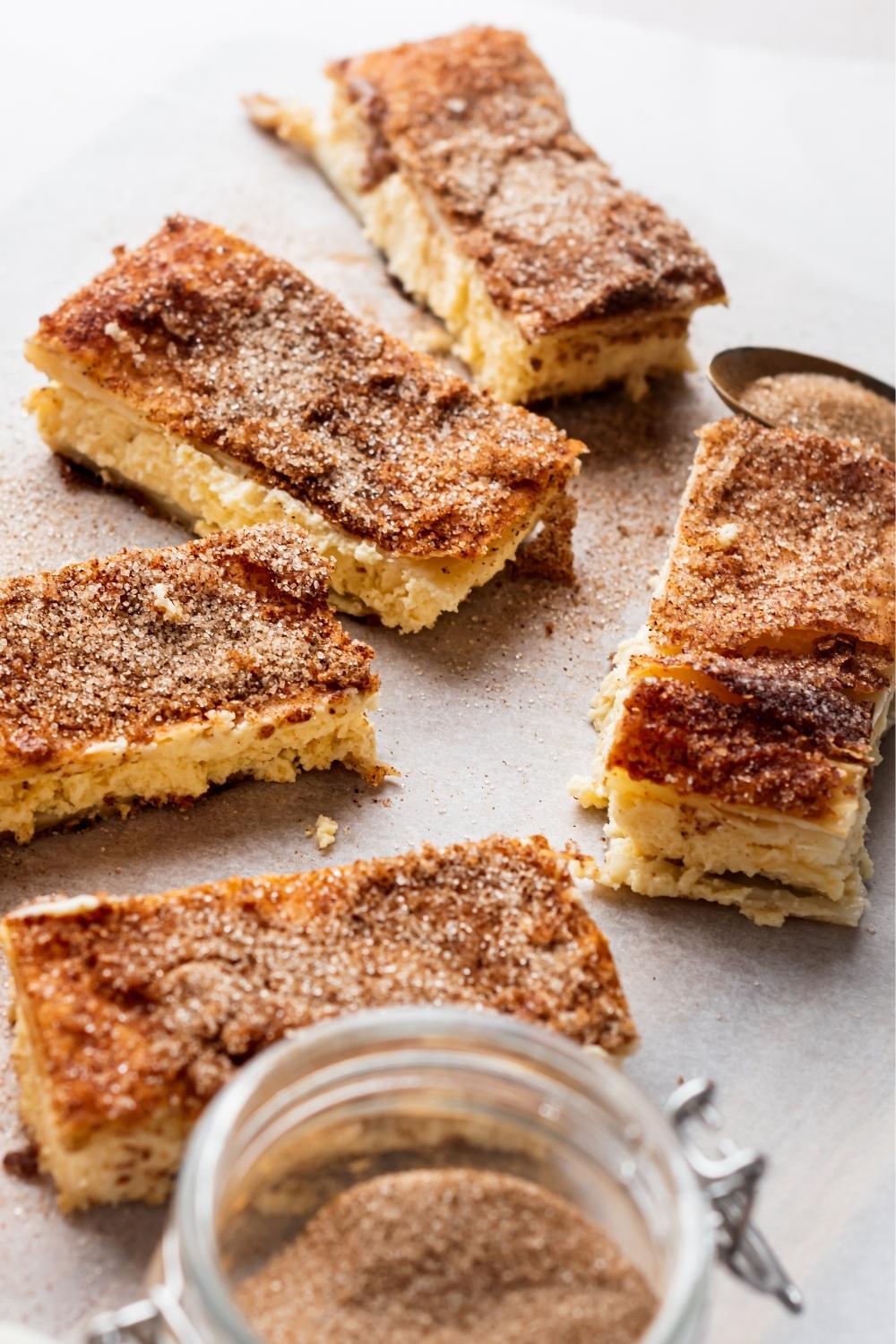 Five churro cheesecake bars sitting on parchment paper topped with cinnamon and sugar. A jar full of cinnamon and sugar sits next to it with a spoon full of cinnamon and sugar.