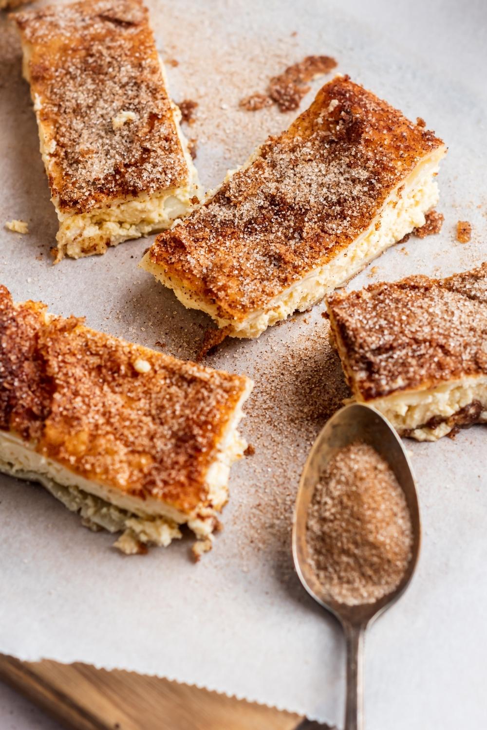 Four churro cheesecake bars on parchment paper. A spoonful of cinnamon and sugar sits next to them.