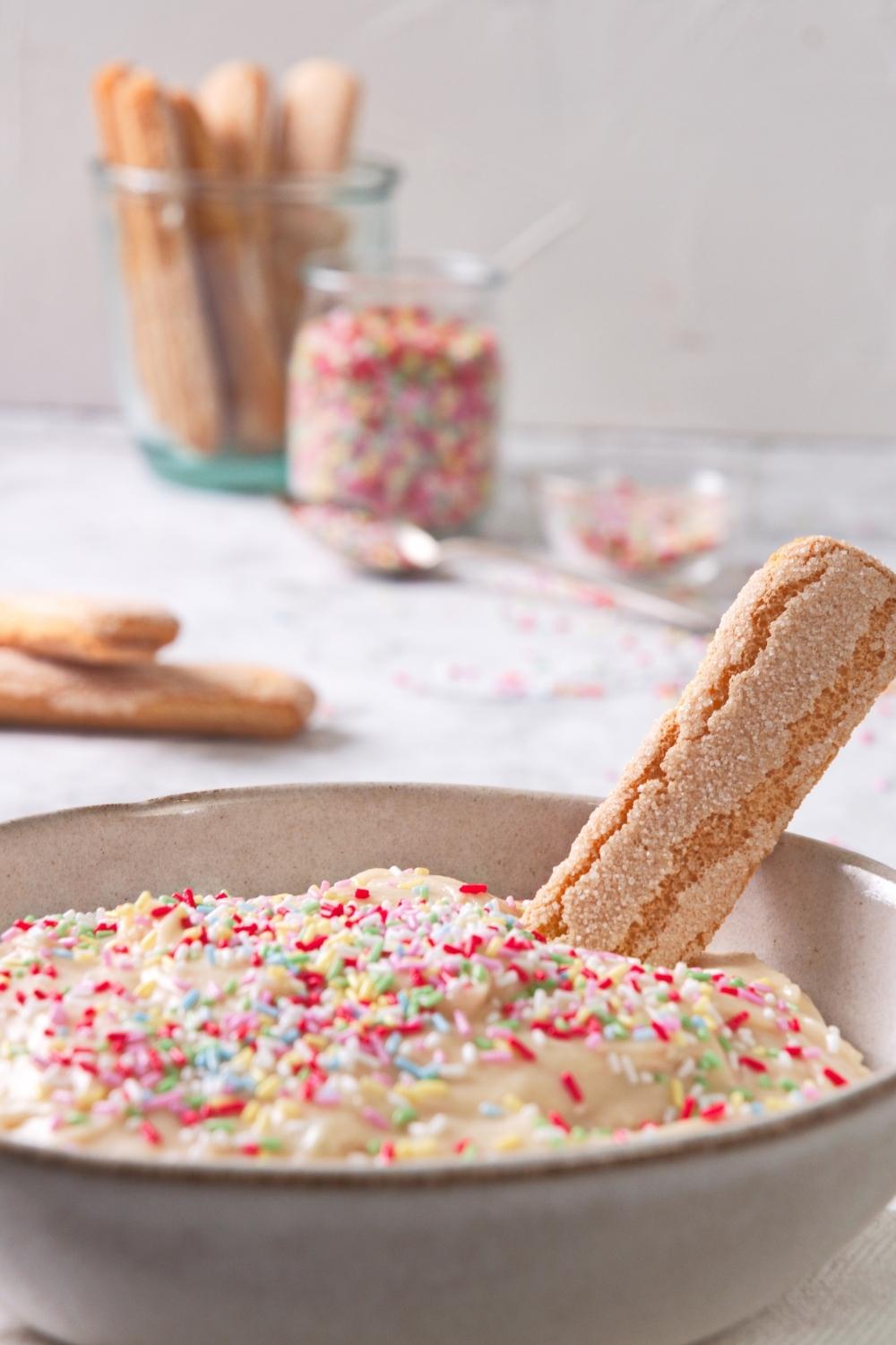 A close-up of a serving bowl containing homemade Dunkaroo dip topped with rainbow sprinkles with a shortbread cookie dipped in it.