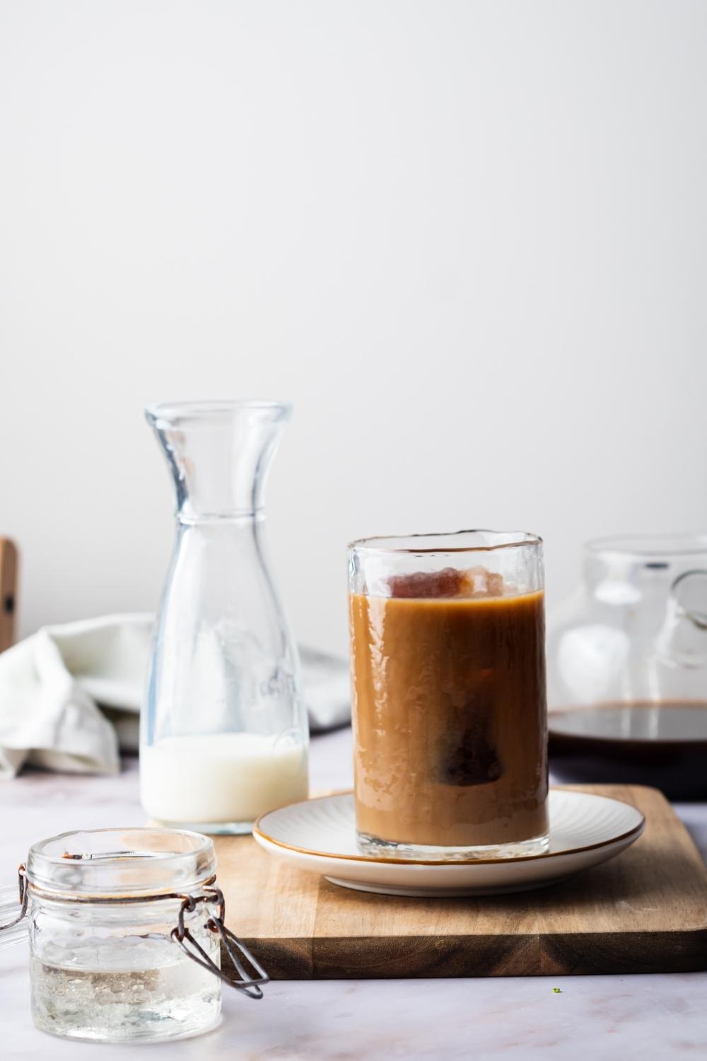 A glass containing iced coffee on a saucer and a pitcher with half and half behind it.