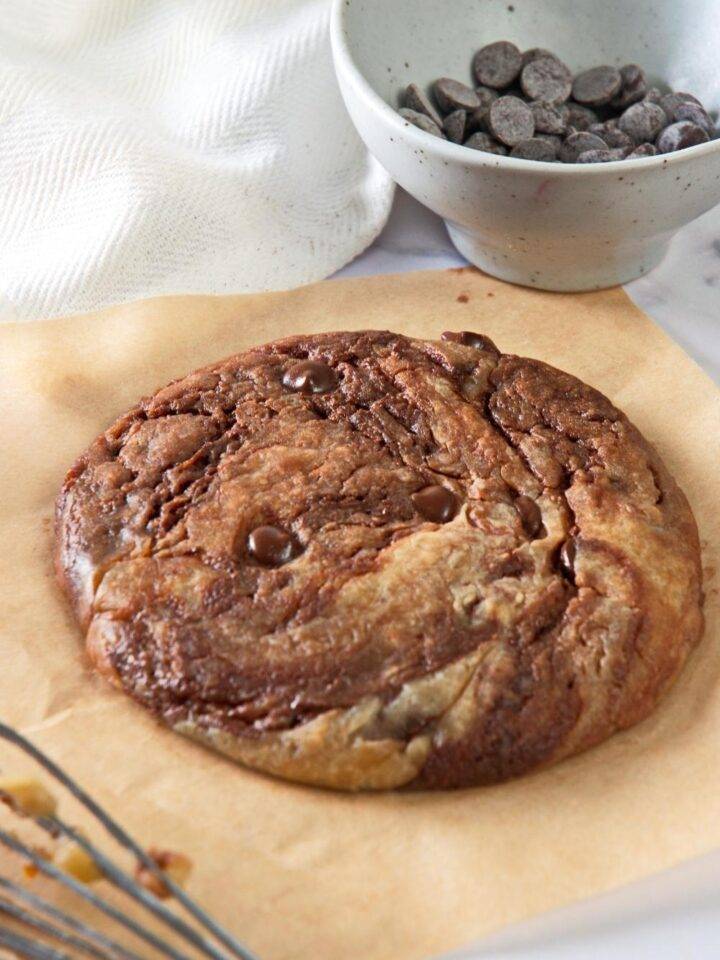 A microwave cookie sits on parchment paper. A whisk lays in front of it and a small bowl with chocolate chips is in the background.